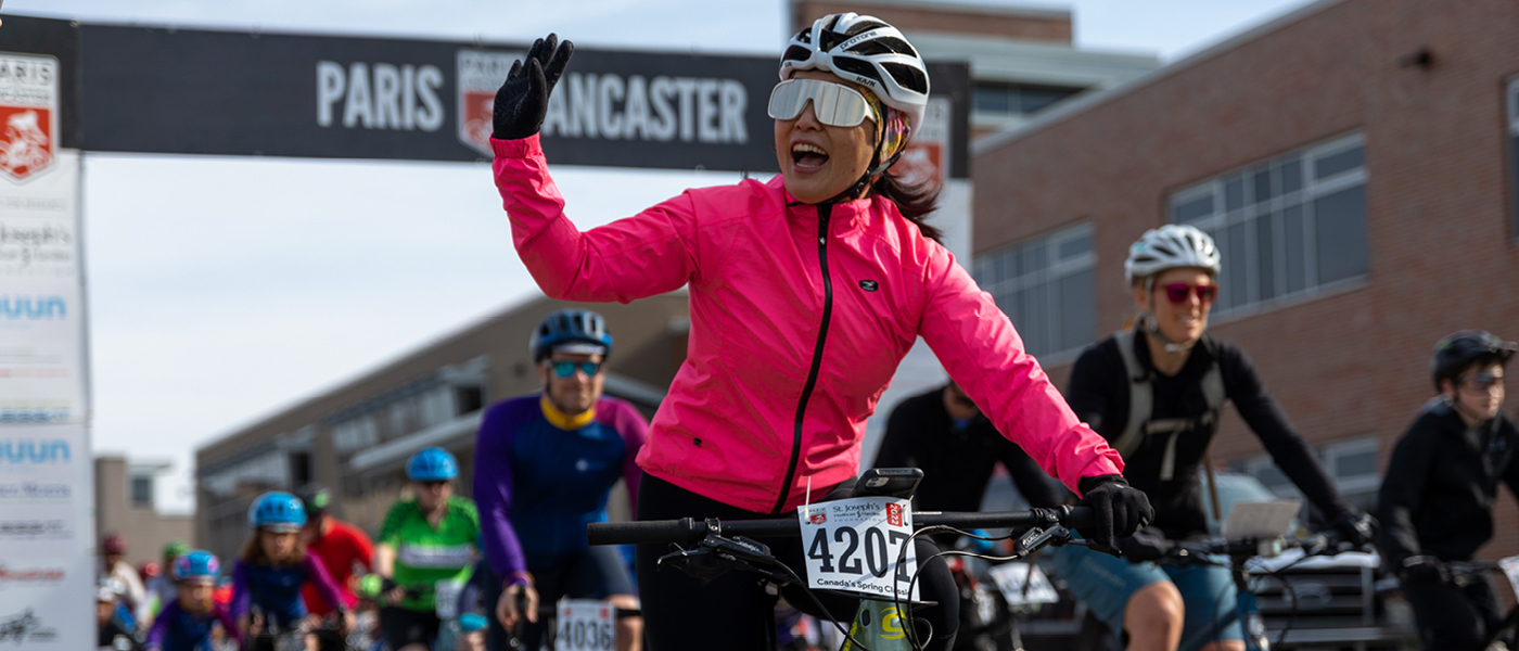 A photo of the start line of the Paris to Ancaster 20km ride. A woman in a pink jackpot waves as she rides by on her bike.