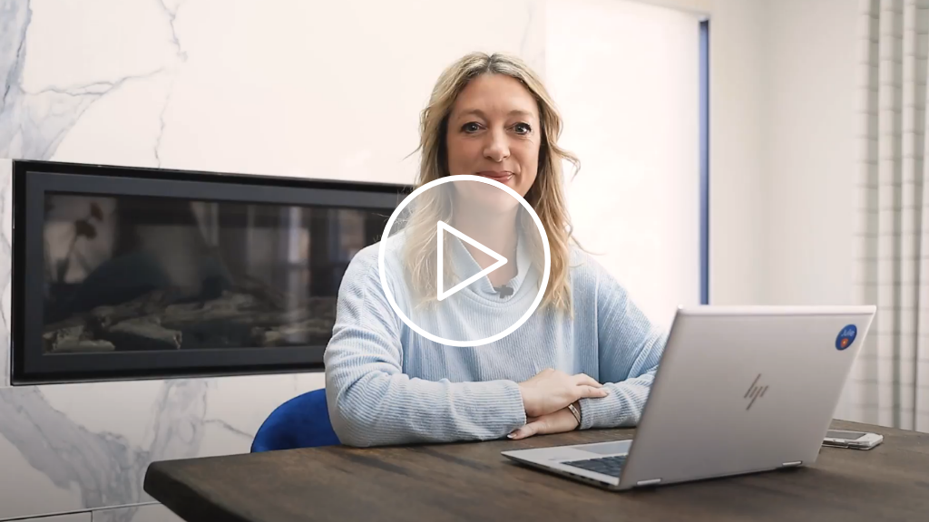 2022 Run for Women Community Champion, Julie Cole, sits at a table in front of a laptop, smiling at the camera with her arms crossed.